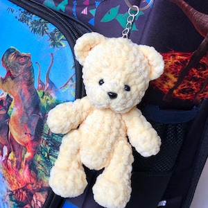 May include: A small, plush, light yellow teddy bear keychain with black eyes and nose. The bear is attached to a silver keyring and is hanging from a backpack with a colorful dinosaur print.