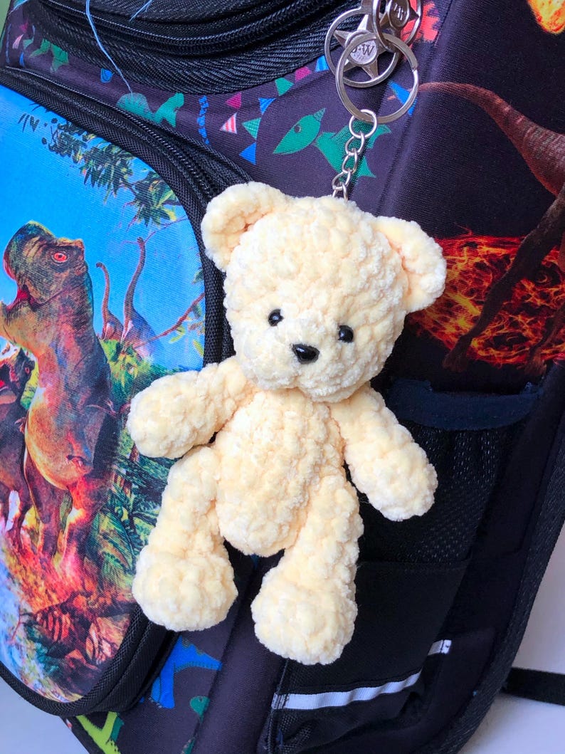 May include: A small, plush, light yellow teddy bear keychain with black eyes and nose. The bear is attached to a silver keyring and is hanging from a backpack with a colorful dinosaur print.