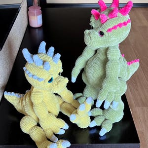 May include: Two handmade crochet dinosaur plush toys. One is yellow with blue accents, the other is green with pink accents. Both have large feet, horns, and friendly expressions. They are on a dark surface.