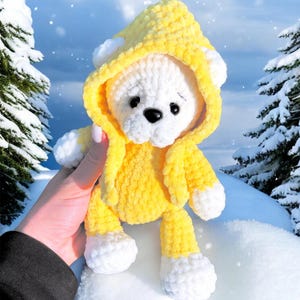 May include: A handmade white and yellow crochet teddy bear wearing a hooded jacket. The bear has a black nose and is set against a snowy background with evergreen trees. The bear's jacket and hood are yellow, and the feet and ears are white.