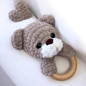 May include: A handmade, plush teddy bear baby rattle in shades of brown and white. The bear has a wooden ring for a handle and a heart-shaped accent. The bear's eyes are black, and the nose is brown.