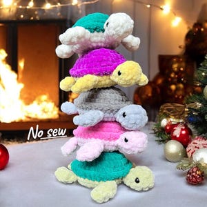 May include: A stack of five crocheted turtles in various colors: teal, white, fuchsia, yellow, gray, and pink. The turtles are arranged in a vertical tower, with the text "No sew" in the lower left corner. The background includes a fireplace and Christmas decorations.