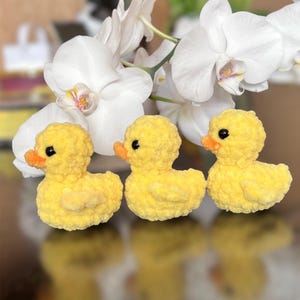 Puede incluir: Tres patitos amarillos de ganchillo hechos a mano con picos naranjas y ojos negros est&aacute;n dispuestos en fila. Los patitos est&aacute;n frente a flores de orqu&iacute;deas blancas. El fondo est&aacute; borroso, con una superficie reflectante.
