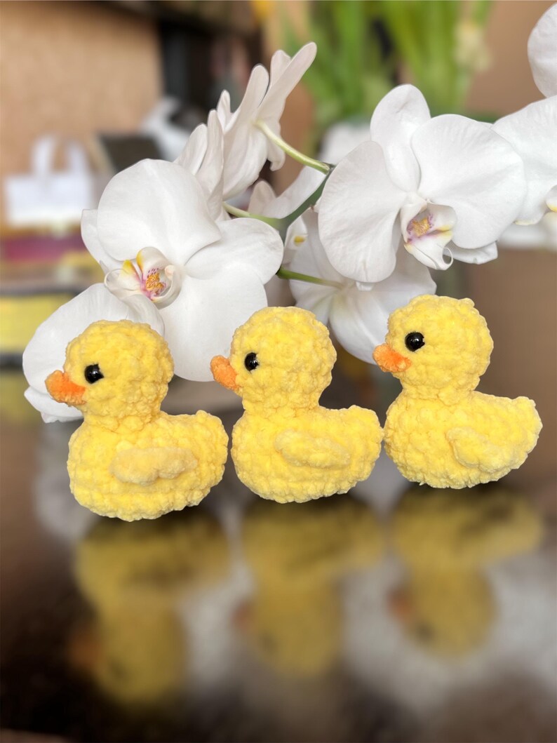 Puede incluir: Tres patitos amarillos de ganchillo hechos a mano con picos naranjas y ojos negros est&aacute;n dispuestos en fila. Los patitos est&aacute;n frente a flores de orqu&iacute;deas blancas. El fondo est&aacute; borroso, con una superficie reflectante.