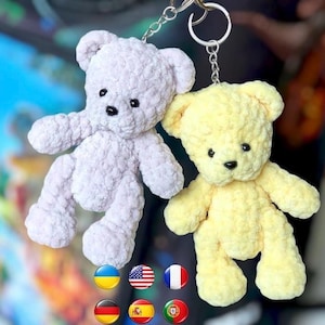 May include: Two handmade teddy bear keychains. One is light purple, the other is yellow. Both have black button eyes and a black stitched nose. The bears are attached to silver keyrings. The background is blurred.