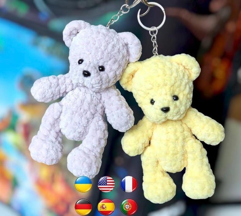 May include: Two handmade teddy bear keychains. One is light purple, the other is yellow. Both have black button eyes and a black stitched nose. The bears are attached to silver keyrings. The background is blurred.