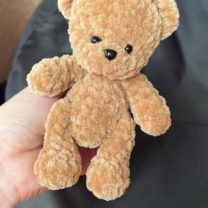 May include: A small, plush teddy bear in a warm tan color. The bear has a soft, textured appearance with black button eyes and a black nose. Its arms and legs are outstretched, giving it a friendly look. A classic children's toy.