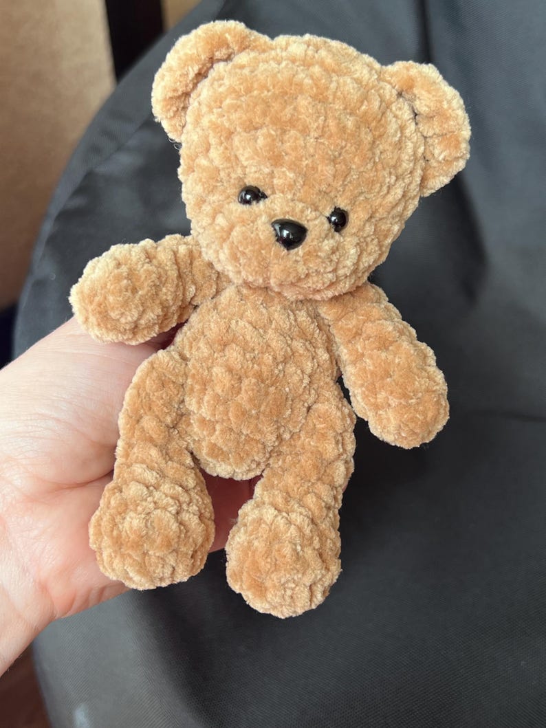 May include: A small, plush teddy bear in a warm tan color. The bear has a soft, textured appearance with black button eyes and a black nose. Its arms and legs are outstretched, giving it a friendly look. A classic children's toy.