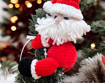Pode incluir: Um enfeite de árvore de Natal feito à mão do Papai Noel. O Papai Noel de pelúcia é vermelho com barba e borda de chapéu brancas. Suas botas pretas têm acabamento branco. O enfeite está em uma árvore de Natal verde com enfeites prateados e azuis.