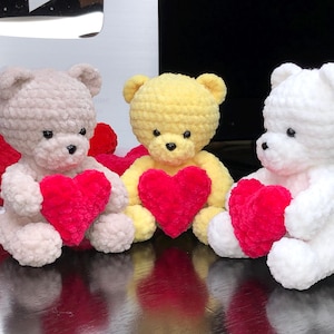 May include: Three crocheted teddy bears holding red hearts. The bears are beige, yellow, and white.