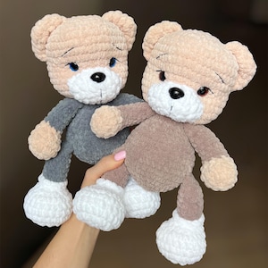 May include: Two handmade crochet teddy bears. One bear is wearing a gray outfit, the other a brown outfit. Both have tan heads, white muzzles, and white feet. The bears have black eyes and stitched features.