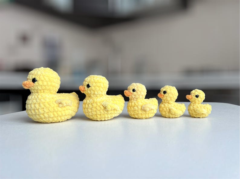 Puede incluir: Una fila de cinco patitos amarillos de ganchillo de diferentes tama&ntilde;os. Cada patito tiene un peque&ntilde;o pico naranja y ojos negros. Los patitos est&aacute;n dispuestos en l&iacute;nea, con el m&aacute;s grande a la izquierda y el m&aacute;s peque&ntilde;o a la derecha.