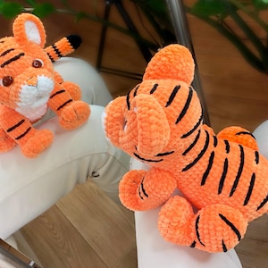 Può includere: Due peluche di tigre arancioni e neri all'uncinetto. Un tigre è seduto con la coda arrotolata attorno al suo corpo. L'altro tigre è sdraiato con la coda distesa dietro di lui.