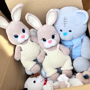 May include: A box filled with handmade crocheted stuffed animals. There are two light brown bunnies with white overalls, a light blue teddy bear with a blue scarf, and a white bunny with black eyes.