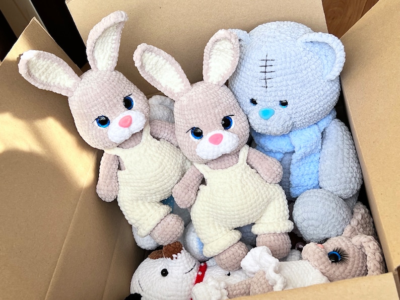 May include: A box filled with handmade crocheted stuffed animals. There are two light brown bunnies with white overalls, a light blue teddy bear with a blue scarf, and a white bunny with black eyes.
