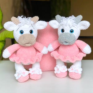 May include: Two handmade crochet cow dolls. The cows have white bodies, pink dresses, and blue eyes. One cow has a gray muzzle and horns, the other has a tan muzzle and horns. They are holding hands in front of a pink pillow.
