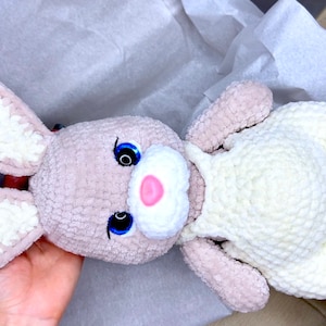 May include: A crocheted plush bunny toy with white and gray fur. The bunny has a white crocheted outfit and blue eyes.