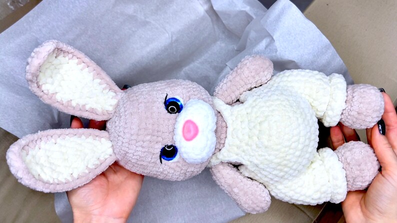 May include: A crocheted plush bunny toy with white and gray fur. The bunny has a white crocheted outfit and blue eyes.