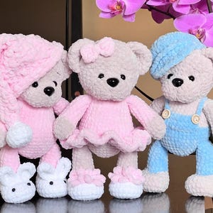 Könnte beinhalten: Drei handgefertigte Häkelteddybären in sanften Pastellfarben. Einer trägt eine rosa Nachtmütze und Häschenschuhe, ein anderer ein rosa Kleid und der dritte ein blaues Outfit und eine blaue Mütze. Jeder Bär hat schwarze Knopfaugen und eine schwarze Nase.