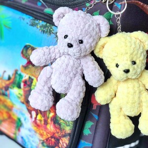 May include: Two handmade teddy bear keychains. One is light purple, the other is yellow. Both have black eyes and noses. They are made of a soft, textured material and are attached to silver keyrings. The background is a backpack with a dinosaur theme.