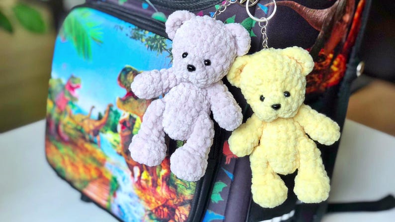 May include: Two handmade teddy bear keychains. One is light purple, the other is yellow. Both have black eyes and noses. They are made of a soft, textured material and are attached to silver keyrings. The background is a backpack with a dinosaur theme.