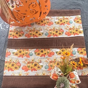 May include: An orange metal pumpkin decoration with cut-out designs, a fall-themed table runner with sunflowers and pumpkins, and a decorative wreath with a bow. The color palette includes orange, brown, and cream.