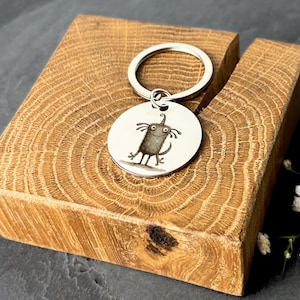 Könnte beinhalten: Ein silberfarbener Schlüsselanhänger mit einem runden Anhänger, der eine skurrile Cartoon-Monster-Illustration zeigt. Der Anhänger ist an einem Schlüsselring befestigt. Der Schlüsselanhänger liegt auf einem Holzblock mit sichtbarer Maserung.