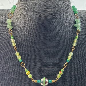 May include: A necklace featuring a gold-tone chain interspersed with various green beads, including translucent round beads, small turquoise seed beads, and a central focal bead with a white, black, and green geometric pattern and green stripes.