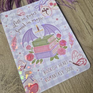 Peut inclure: Un autocollant rectangulaire sur le thème de la fantaisie. Il présente une pile de livres avec des ailes violettes et une épée, entourée de roses, de papillons et du texte "Lost in pages found in magic" et "Bookish Heart Romantasy Soul".