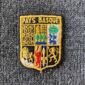 Peut inclure: Écusson brodé avec le texte "PAYS BASQUE" et un blason coloré. Le patch a une bordure noire avec une garniture dorée et est posé sur un fond en tissu tricoté gris.