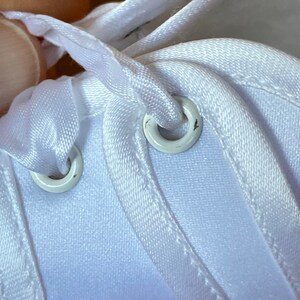 May include: Close-up of white shoelace eyelets on a white fabric shoe. The eyelets are metal and have a white finish.