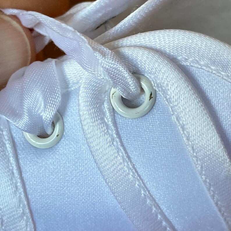 May include: Close-up of white shoelace eyelets on a white fabric shoe. The eyelets are metal and have a white finish.