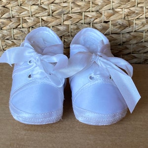 May include: A pair of white satin baby shoes with white satin ribbon laces. The shoes have a white trim around the sole.