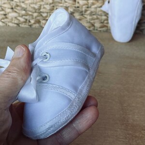 May include: White baby shoes with laces and a white satin bow. The shoes have a white sole and are made of a soft, white fabric.