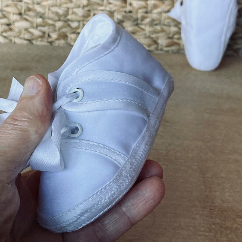 May include: White baby shoes with laces and a white satin bow. The shoes have a white sole and are made of a soft, white fabric.