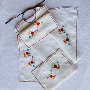 Pode incluir: Um conjunto de babete e guardanapos de linho branco com bordados florais. O babete tem uma fita castanha, e os guardanapos apresentam flores vermelhas, amarelas e azuis com caules verdes. As bordas são aparadas com costuras castanhas.