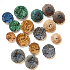 Op de afbeelding: Een verzameling van vijftien ronde knopen in verschillende maten en kleuren, waaronder tinten blauw, groen, bruin en beige. Elke knoop heeft een getextureerd oppervlak met een opvallend centraal balkontwerp en twee kleine gaatjes.