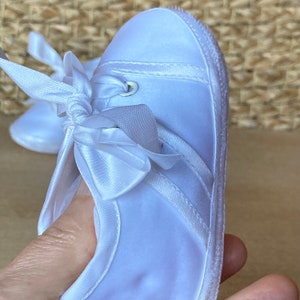 May include: White satin baby shoes with a white satin bow and a white eyelet detail. The shoes have a white sole.