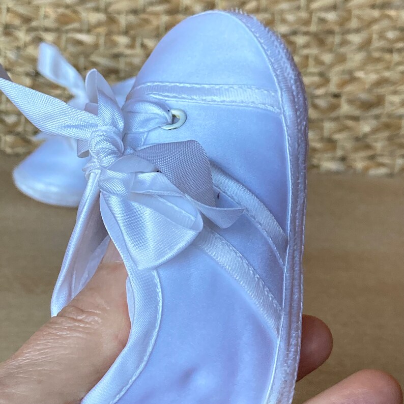May include: White satin baby shoes with a white satin bow and a white eyelet detail. The shoes have a white sole.