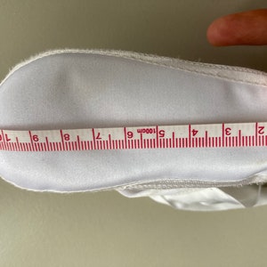 May include: White baby shoe with a white satin ribbon and a white fabric sole. The shoe is being measured with a tape measure that reads 10cm.