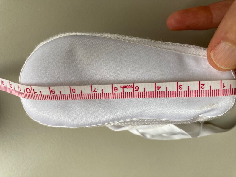 May include: White baby shoe with a white satin ribbon and a white fabric sole. The shoe is being measured with a tape measure that reads 10cm.