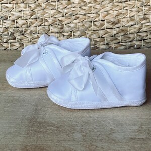 May include: A pair of white satin baby shoes with satin bows and a white sole.