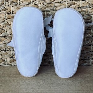 May include: A pair of white baby shoes with satin bows. The shoes are made of soft, breathable fabric and have a flexible sole.