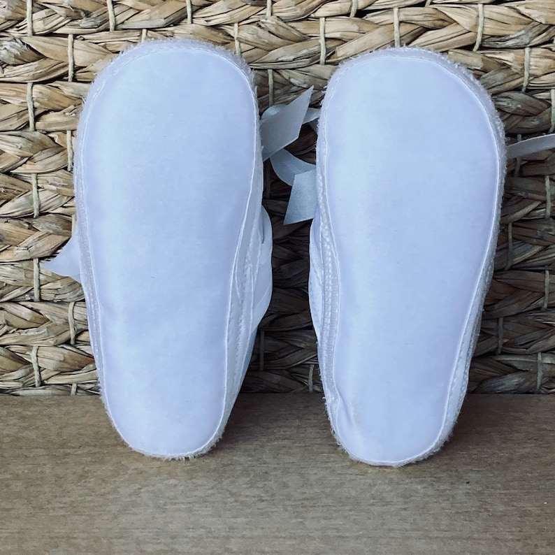 May include: A pair of white baby shoes with satin bows. The shoes are made of soft, breathable fabric and have a flexible sole.