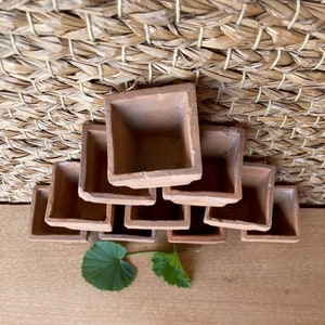 Op de afbeelding: Een stapel van acht vierkante terracotta bloempotten, gerangschikt in een piramidevorm. De potten zijn ongeglazuurd en hebben een natuurlijke bruine kleur.