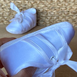 May include: A pair of white satin baby shoes with a bow and lace-up closure. The shoes are made of soft, comfortable material and have a delicate design.