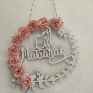 Pode incluir: Uma coroa de madeira branca com um design floral e as palavras "Eid Mubarak" em itálico. A coroa é decorada com flores de tecido rosa.