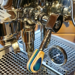 Könnte beinhalten: Nahaufnahme einer silbernen Espressomaschine mit einem Holzhebel. Der Hebel ist mehrfarbig mit blauen, grünen und braunen Streifen. Die Maschine hat ein Manometer mit schwarzem Zifferblatt und weißen Markierungen. Das Manometer zeigt "8 bar" und "12 bar" an. Die Maschine trägt ein Etikett mit der Aufschrift "Rocket Espresso Milano Mozzafiato Evo R".