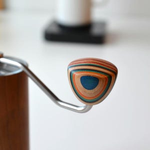 Könnte beinhalten: Nahaufnahme eines Kaffeemühlen-Griffs. Der Griff besteht aus geschichtetem Holz in verschiedenen Farben, darunter Blau, Orange und Braun. Der Griff ist dreieckig geformt und an einem Edelstahlarm befestigt. Das Mühlengehäuse ist braun.