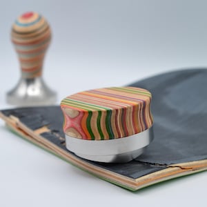 Könnte beinhalten: Ein bunter, gestreifter, hölzerner Kaffeestamper mit silberner Basis, der auf einem Skateboard-Deck liegt.
